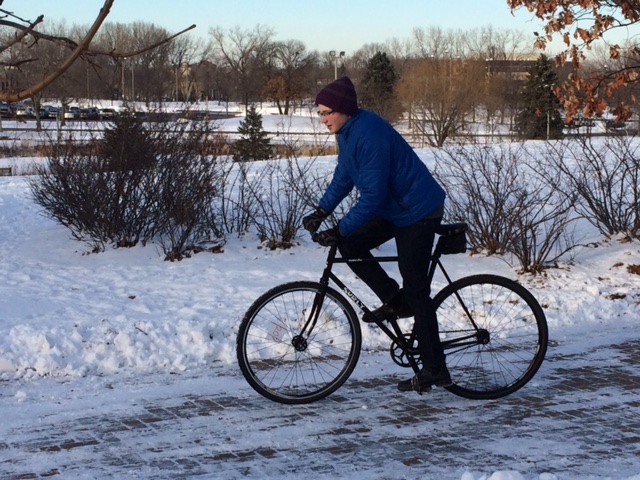 Left side view of a winter cyclist on a black Surly bike, riding on a icy brick sidewalk
