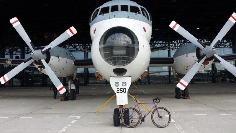 Right side view of a purple Surly bike, parked in an aircraft hanger against the front wheels of a plane facing forward