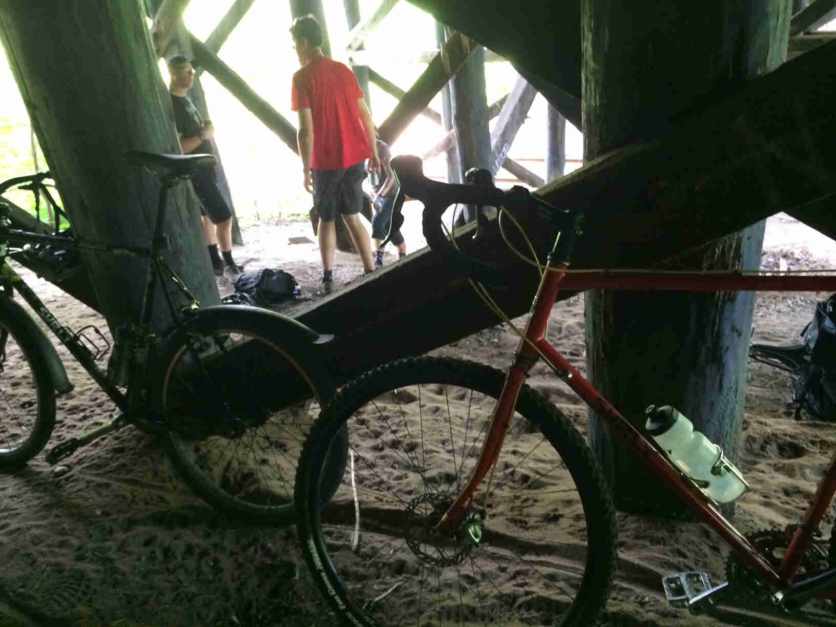 Left side view of 2 Surly bikes, leaning on a post single-filed under a pier, with 3 people standing near