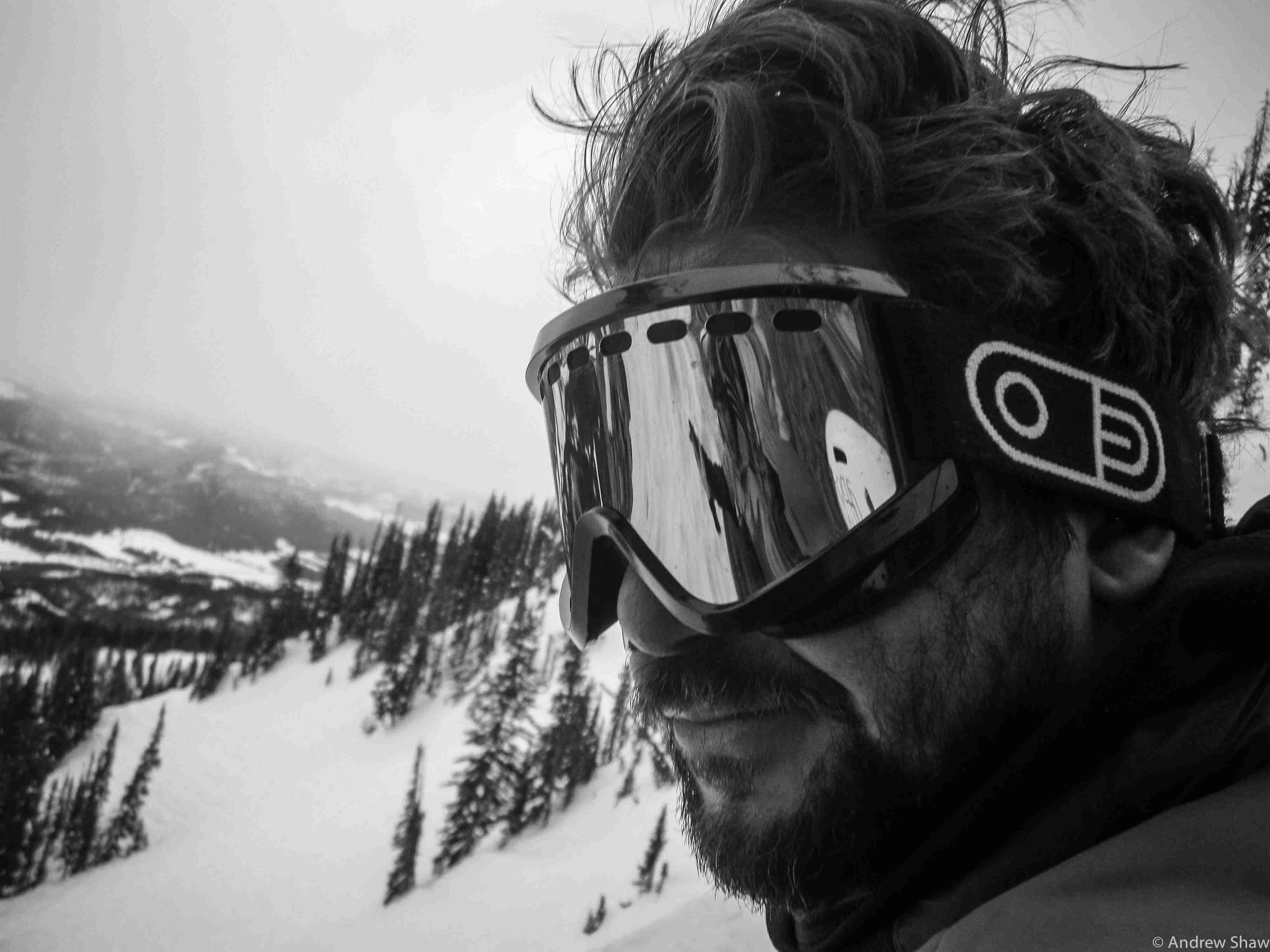 Left side headshot of a person wearing ski goggles, with a snow covered mountain hill behind them - black & white image
