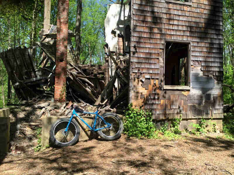 Left side view of a blue Surly fat bike, parked on dirt, in front of a crumbling wood building in the forest