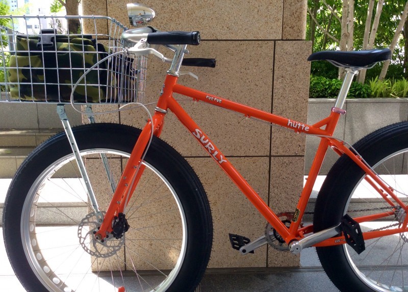 Left side view of an orange Surly Pugsley fat bike with a front basket, parked against a stone block pillar