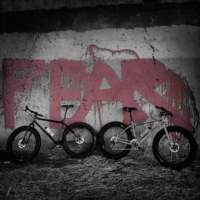 Side view of 2 Surly Pugsley fat bikes, facing opposite directions, against a concrete wall with Graffiti