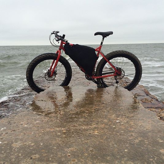 Left side view of a red Surly Pugsley bike with a frame pack, parked across a low cement pier on the ocean
