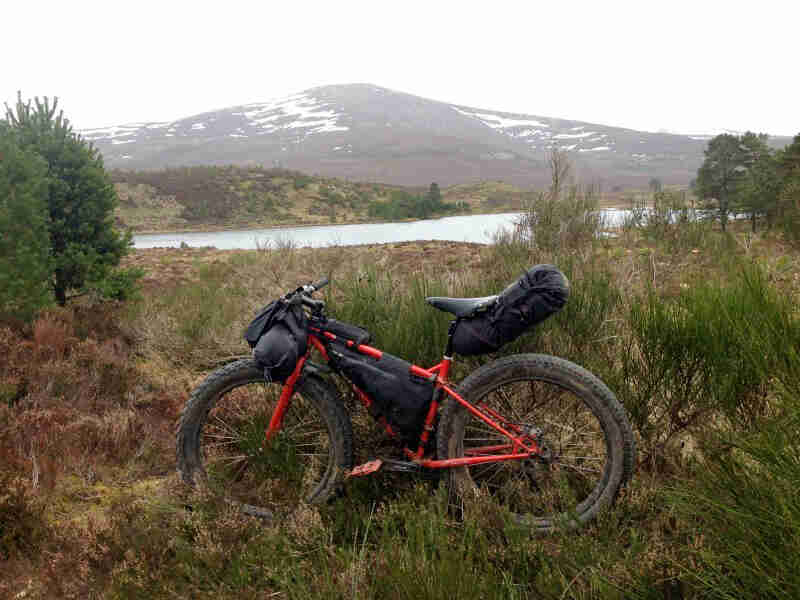 Left side view of a red Surly Pugsley fat bike with gear packs, parked on grass, with a lake and mountain in background