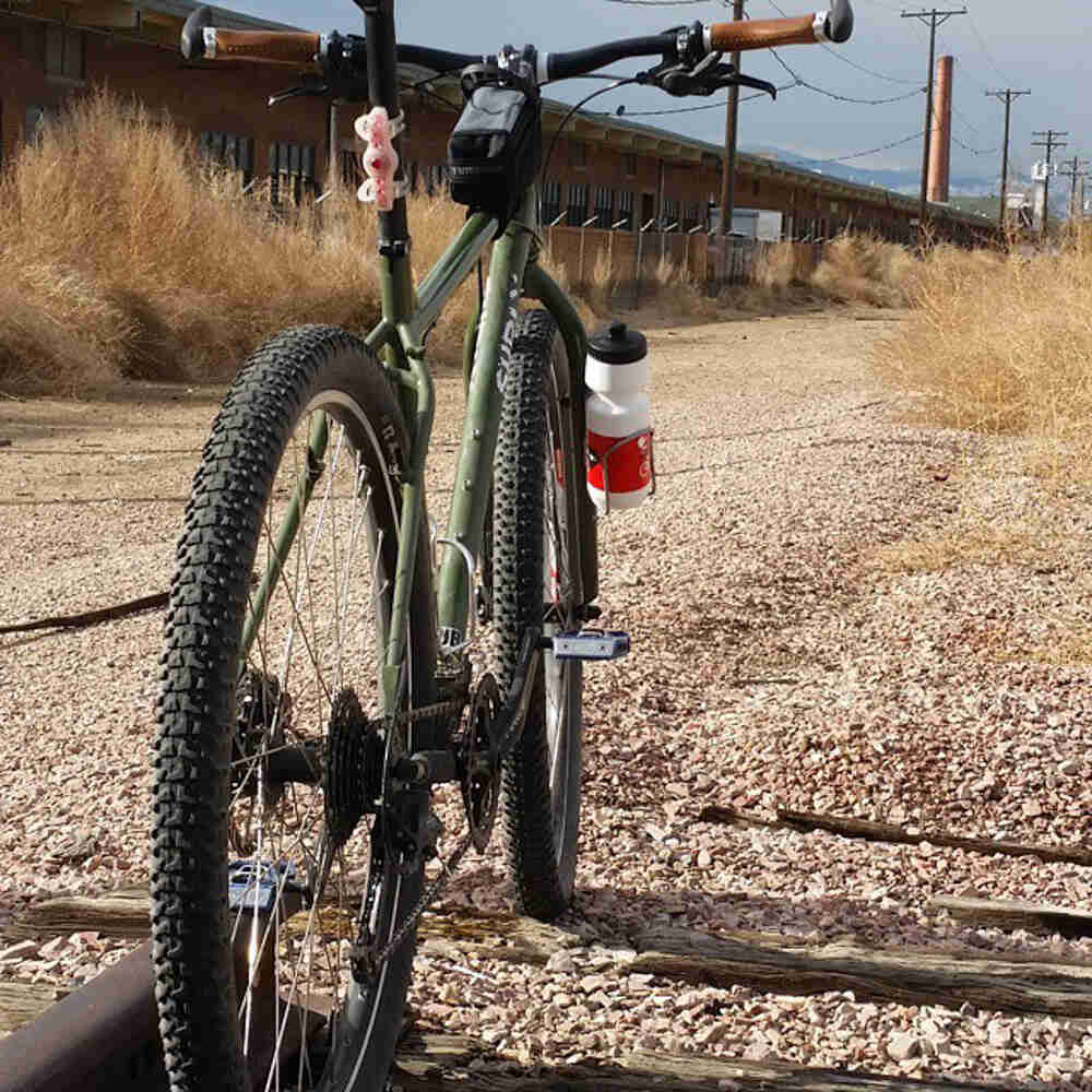 Rear view of an olive green Surly bike, parked in a gravel field, with long brick building to the left