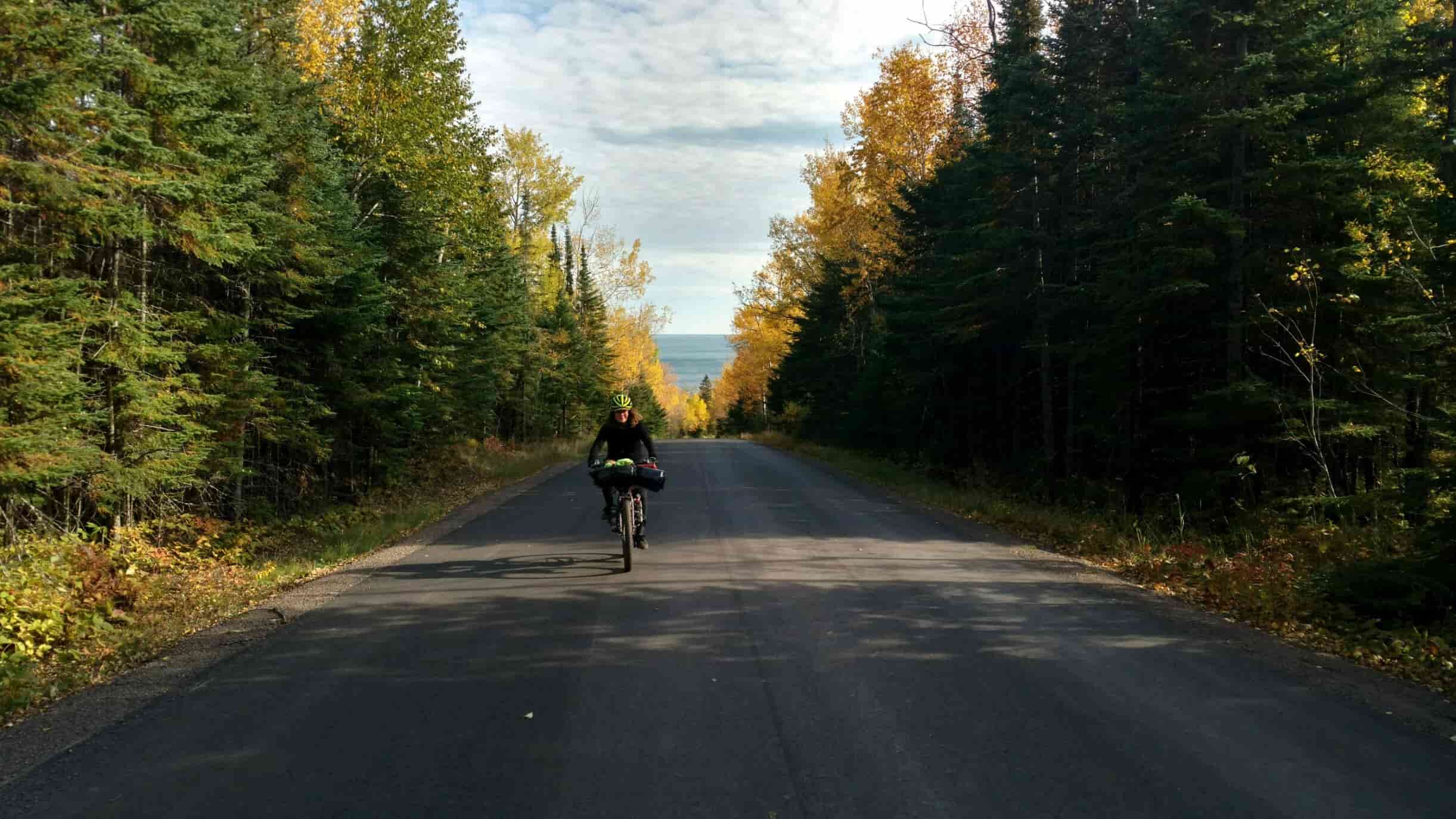 Front view of a cyclist riding down the middle of a paved road with pine trees on both side