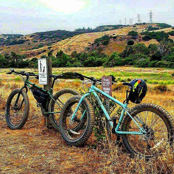 Left side view of 2 Surly bikes, parked in grass against trail signs, with a grassy field and hills in the background