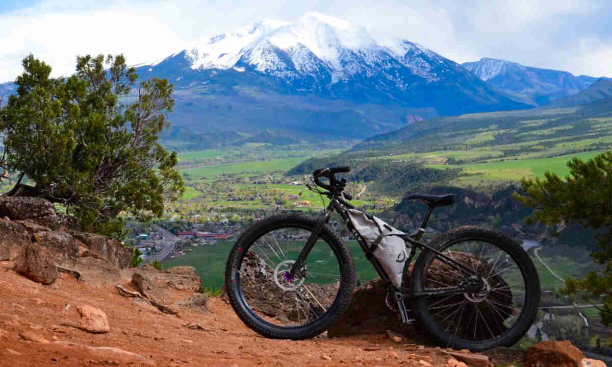 Left side view of a black Surly fat bike, parked on a red rock hill, a with snow capped mountain in the background