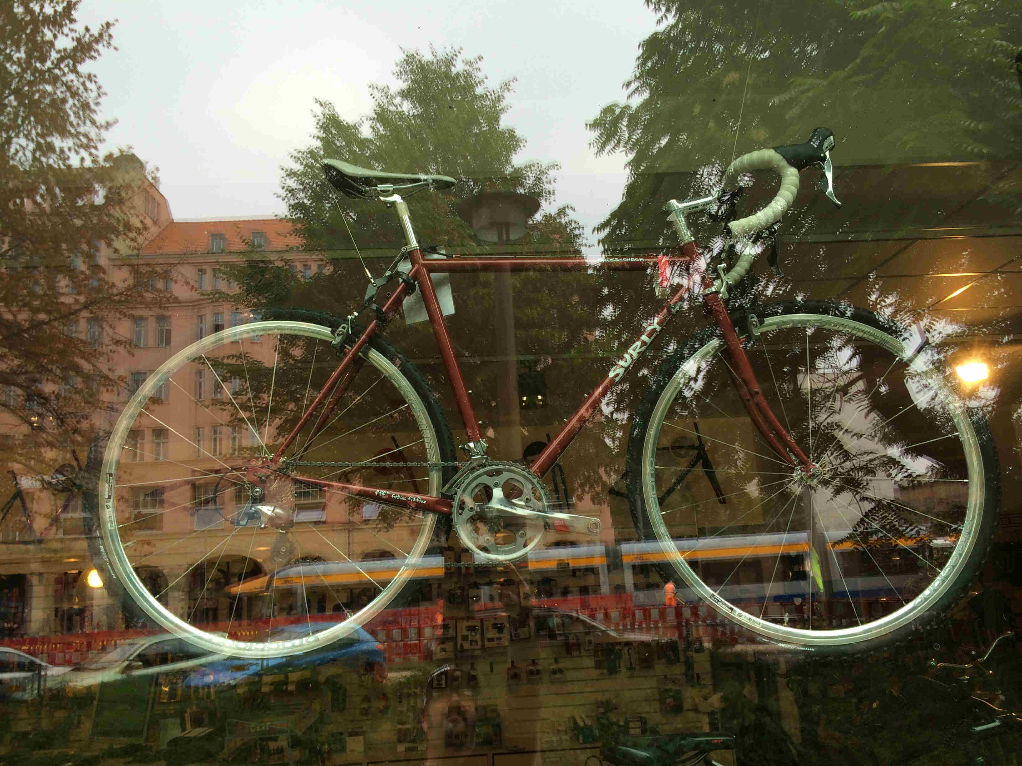 Right side view of a copper Surly Cross Check bike, hanging behind a bike shop's store front window