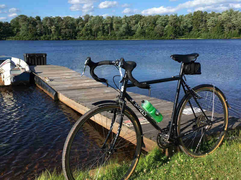 Left side view of a Surly bike, black, on the bank of a lake, in front of a wood dock, with pine trees in the background