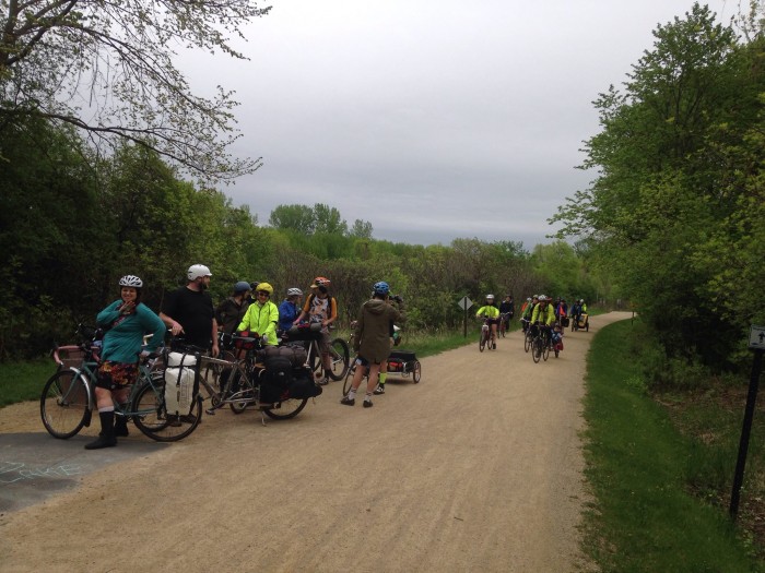 A group of cyclists, riding or standing with their bikes, on a gravel road with trees on both sides