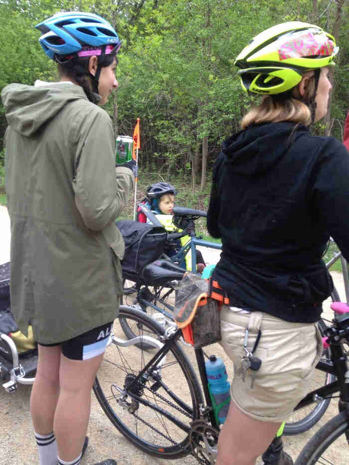 Rear view of 2 cyclists, standing with their bikes and a child in rear bike seat next to them, with trees in background