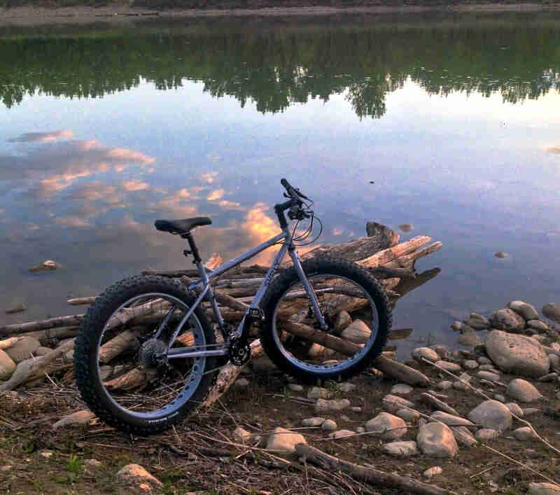 Right side view of a Surly fat bike, gray, parked on a rocky lakeshore, leaning against a pile of logs