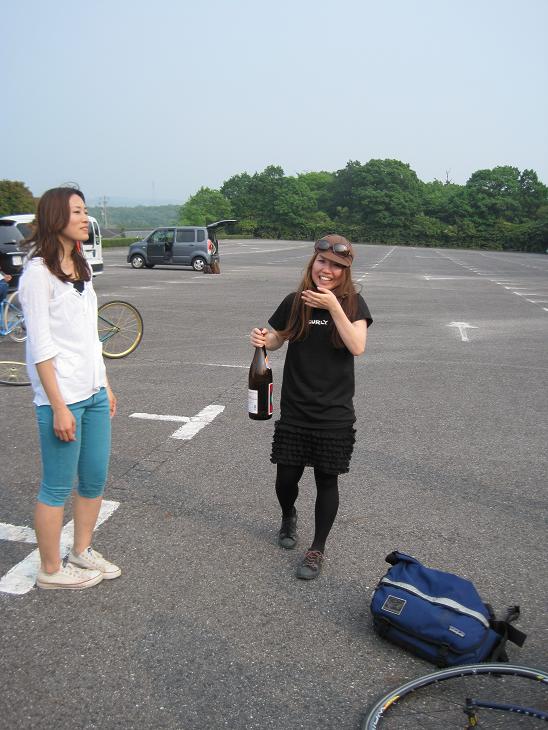 Front view of a person, wearing a Surly t-shirt and holding a large bottle of sake next to someone, on a parking lot