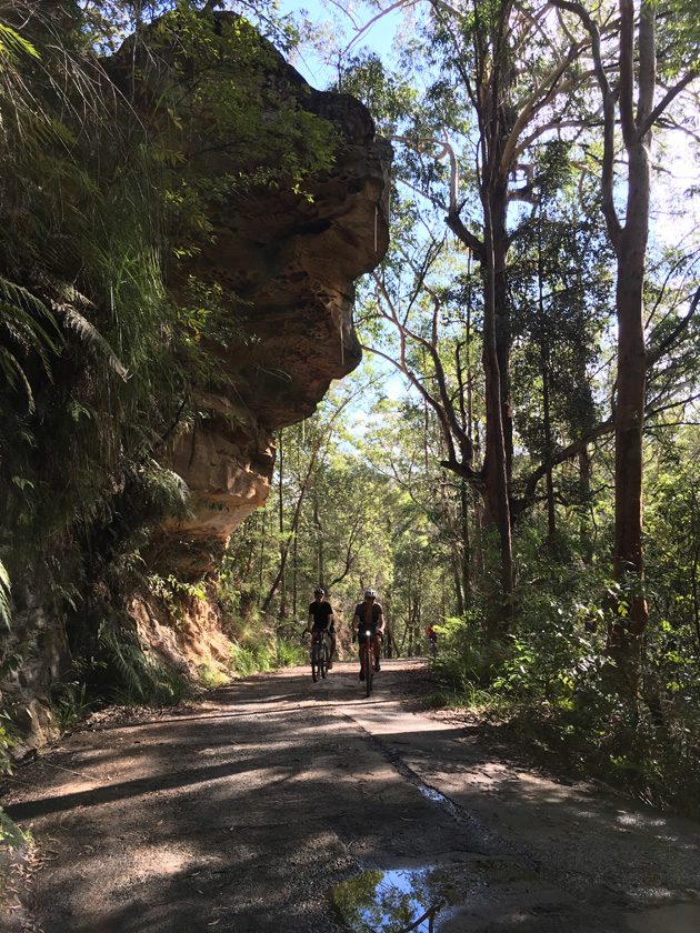 Two cyclist ride side by side down a dirt trail with a rock cliff leaning over and trees shown in the background