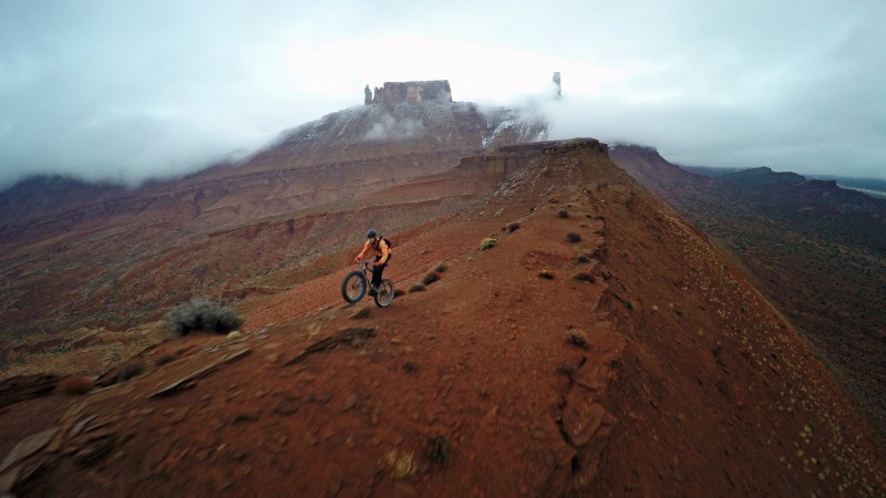 Front, left side view of a cyclist, riding a Surly fat bike down  mountain ridge, with a fog covered castle behind them