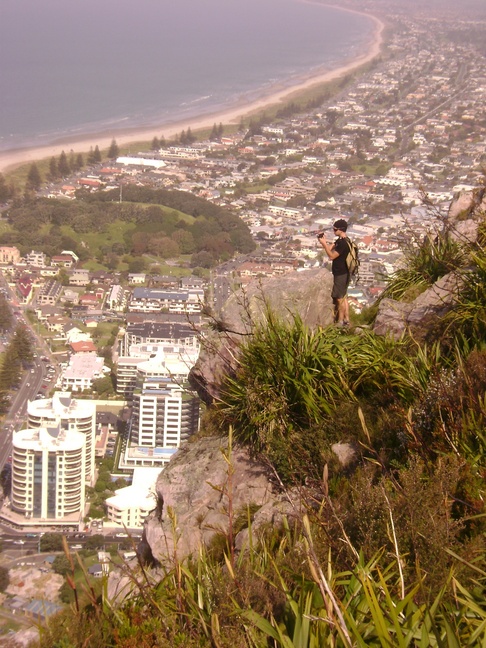 Left side view of a person standing on a cliff edge, high above a city that's along an ocean shore