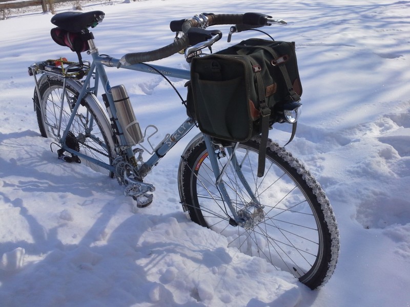 Right side view of a light blue Surly Trucker Deluxe bike with a front pack, standing in a field of deep snow