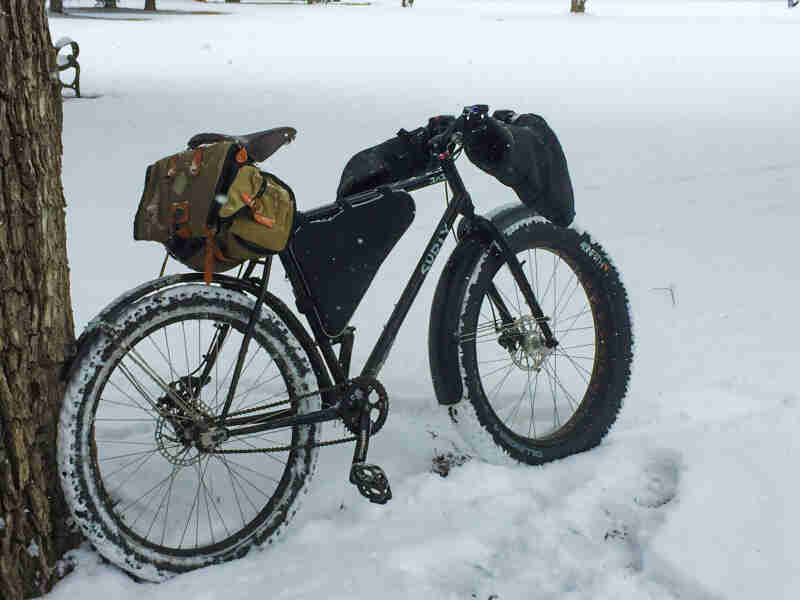 Right side view of a black Surly fat bike, loaded with packs, parked in a snow covered field