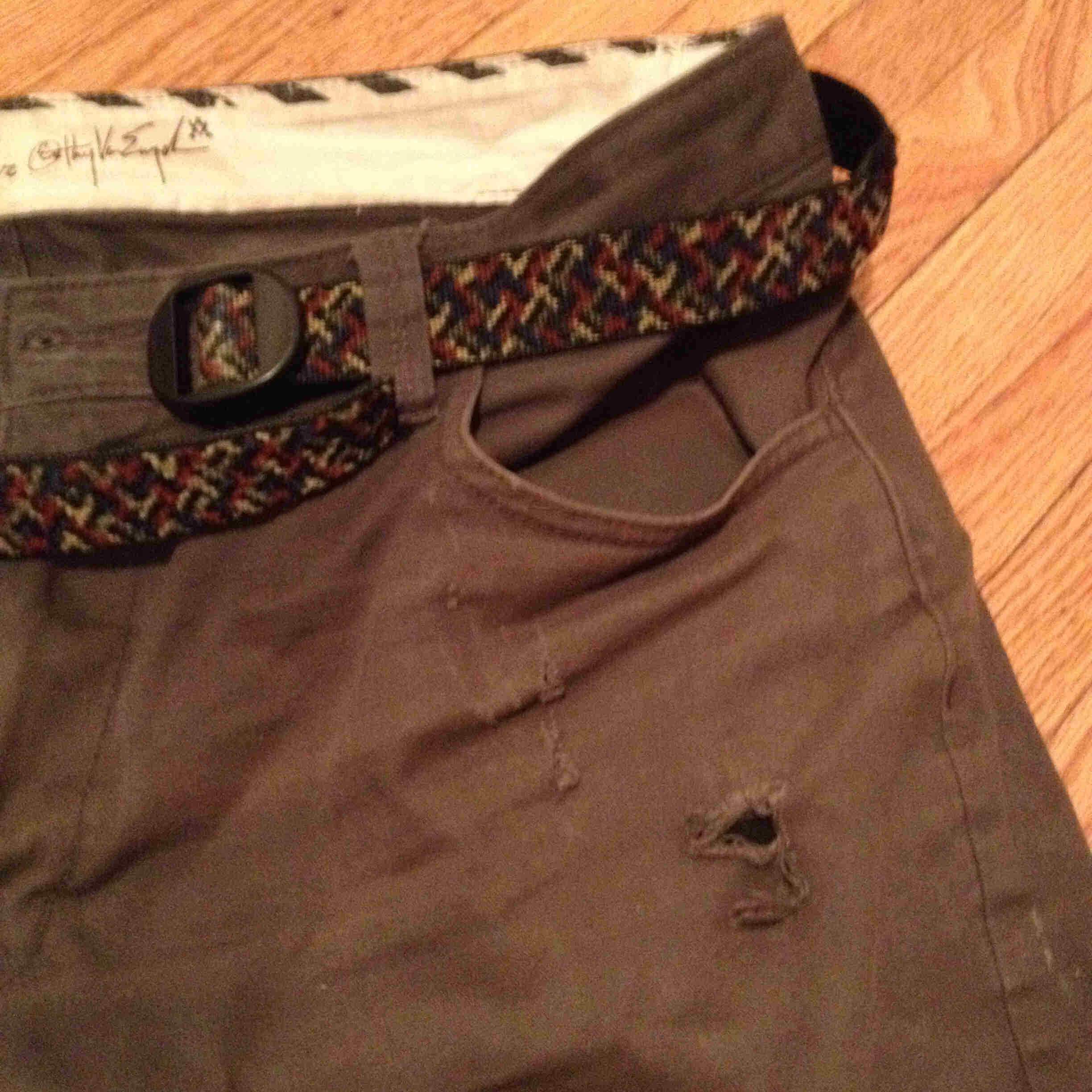 Downward, front, upper left side view of a pair of brown shorts, with a hole in the left pocket