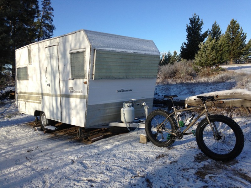 Right side view of a tan Surly Moonlander fat bike, parked in snow, with the back wheel at the hitch on a towable camper