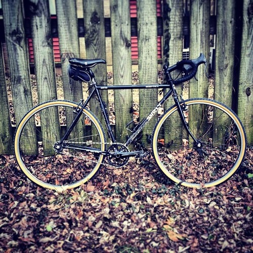 Right side view of a black Surly Cross Check bike, leaning against a wood fence wall, with leaves on the ground