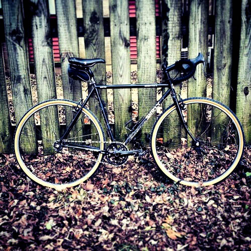 Right side view of a black Surly Cross Check bike, parked on leaves and leaning against a wood fence wall