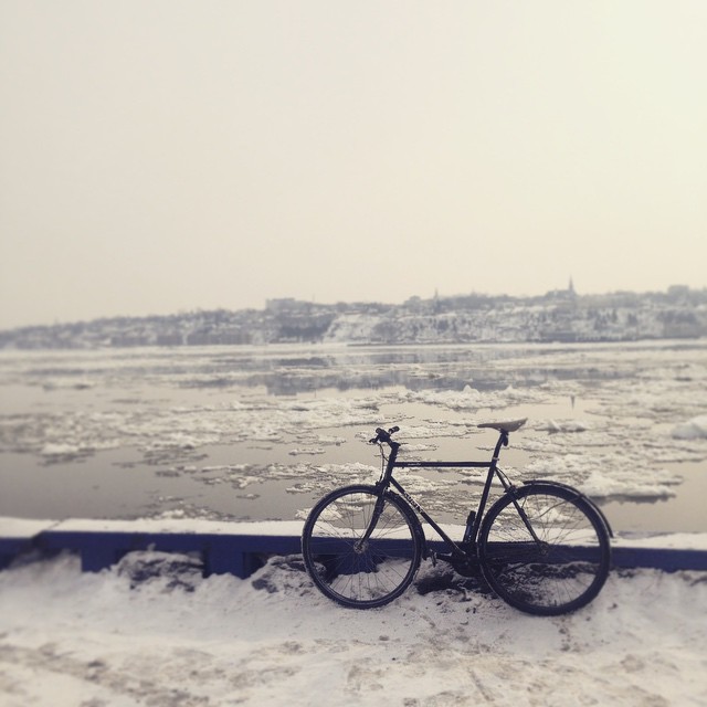 Left side view of a black Surly Steamroller bike, parked on snow against a short wall, with icy waters in the background