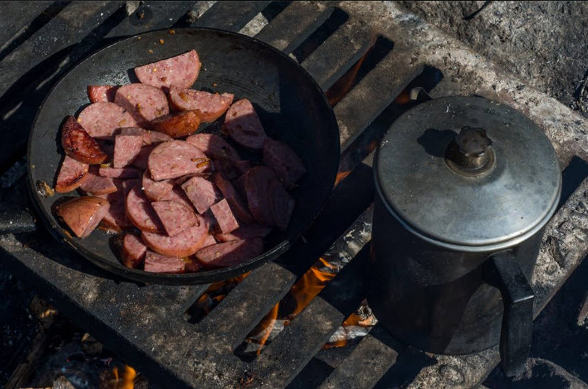 Downward view of a pan of cut up sausage and a coffee pot, sitting on top of a grate over a fire