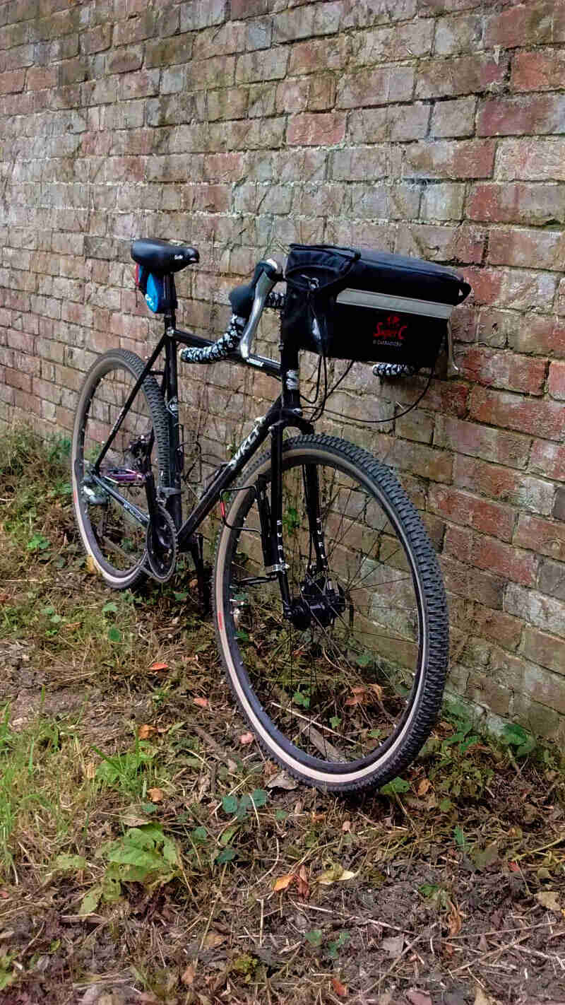 Front, right side view of a black Surly straggler bike with a handlebar pack, leaning against a brick wall