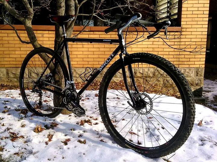 Right side view of a black Surly Straggler bike, leaning against a tree, on top of snow in front of a short brick wall