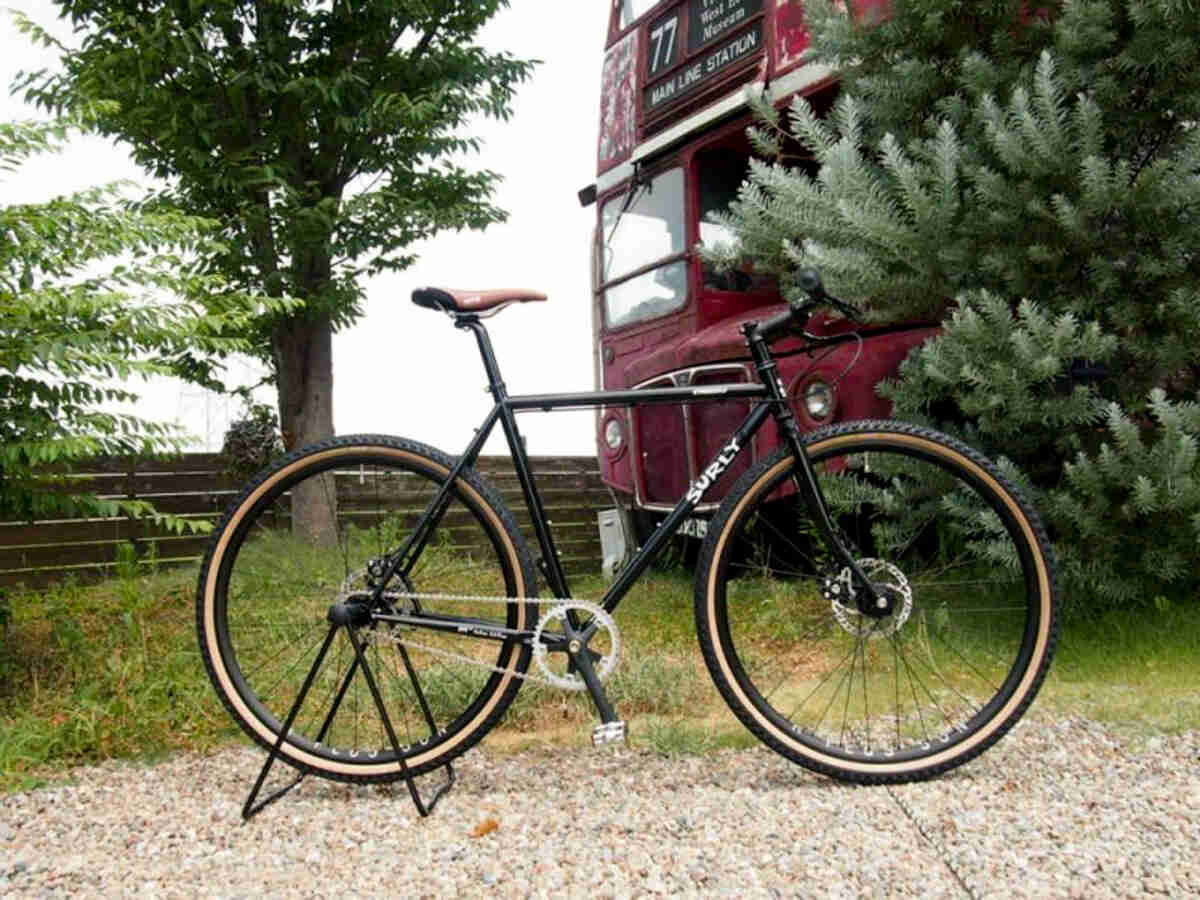 Right side view of a black Surly Straggler bike, parked in front of a double decker bus peaking out from a tree