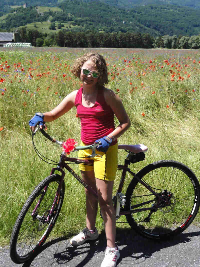 Left side view of a purple Surly Straggler bike with a cyclist standing over, in front of a grass field with flowers