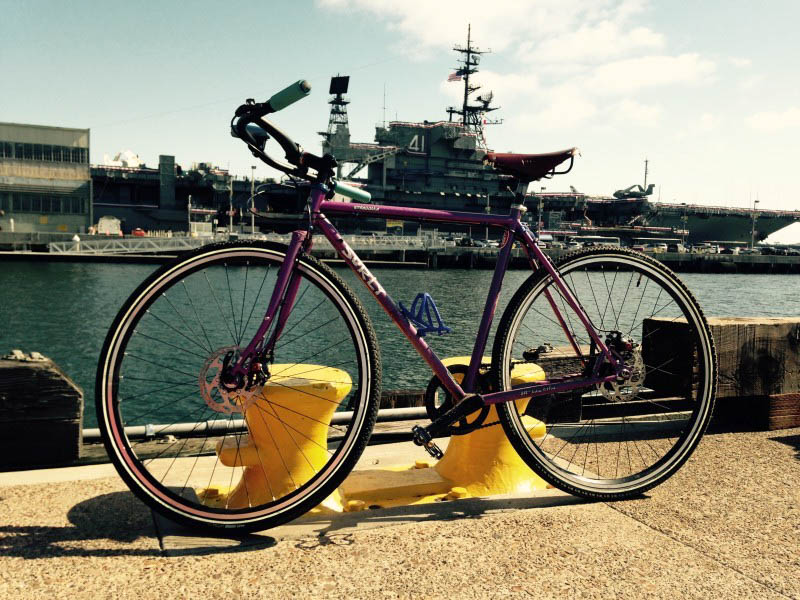 Left side view of a purple Surly Straggler bike, on a concrete dock at a port, with an aircraft carrier in background