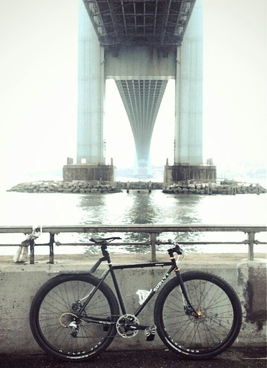 Right side view of a black Surly Straggler bike, against a short concrete wall with a waterway behind, under a bridge