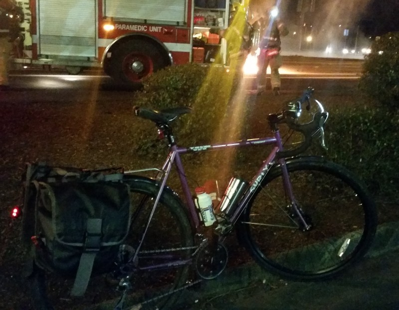 Right side view of a purple Surly Straggler bike with gear, parked on a curb at night, with a firetruck in background