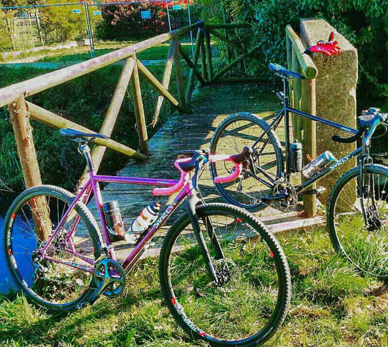 Front right side view of 2 Surly bikes, in front of a wood bridge, over a stream 
