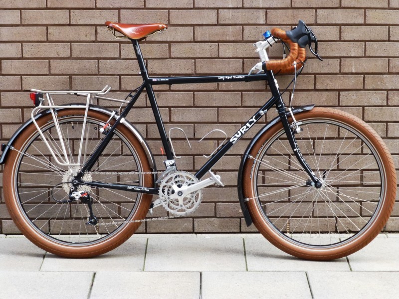 Right side view of a black Surly Long Haul Trucker bike with brown tires, on a sidewalk, leaning against a brick wall