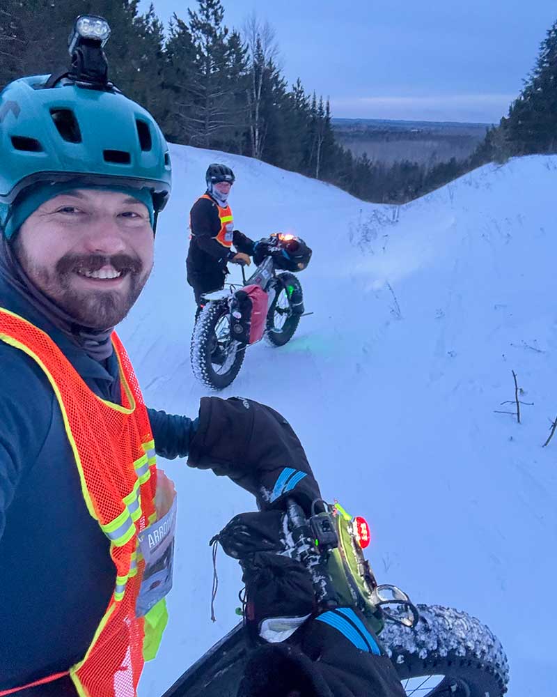 Alex smiling taking selfie on trail with Ian in background atop hill