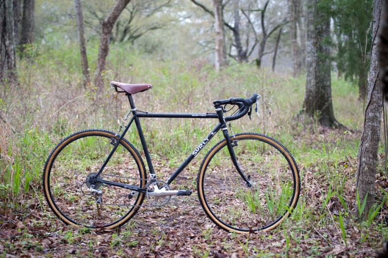 Right side view of a black Surly Cross Check bike, parked on grass and leaves, with weeds and trees in the background