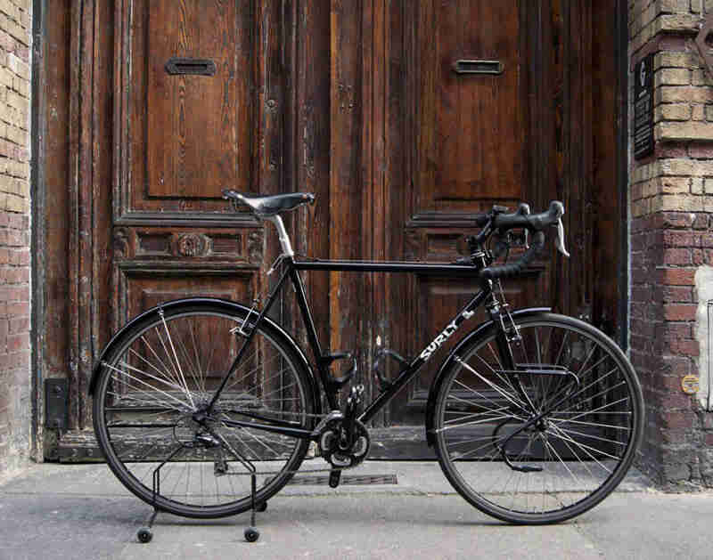 Right profile of a black Surly road bike on a front step, in front of a building with 2 wooden doors