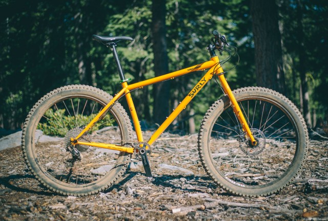 Surly Karate Monkey 27.5+ bike - yellow - parked on sticks with a forest in the background - right profile view