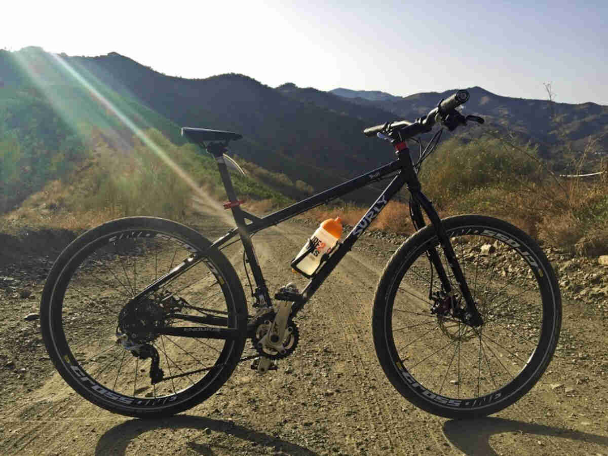 Right side view of a black Surly Troll bike, parked across a gravel road, with tree covered mountains in the background