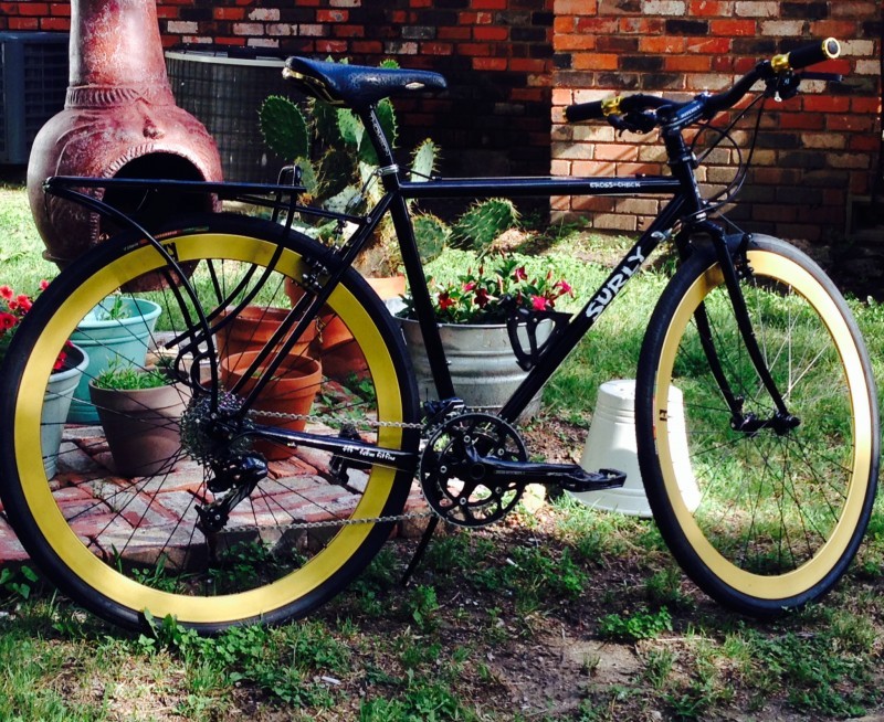 Right side view of a black Surly Cross Check bike with yellow rims, parked in front of potted plants, on a grass yard