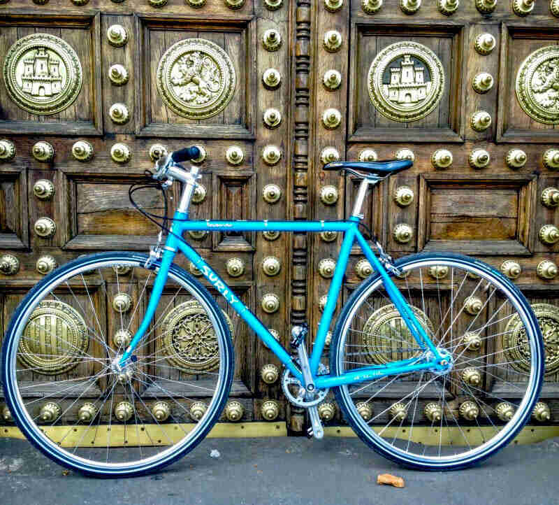 Left side view of a turquoise Surly Steamroller bike, parked in front of 2 large wood doors with gold decorative hardware