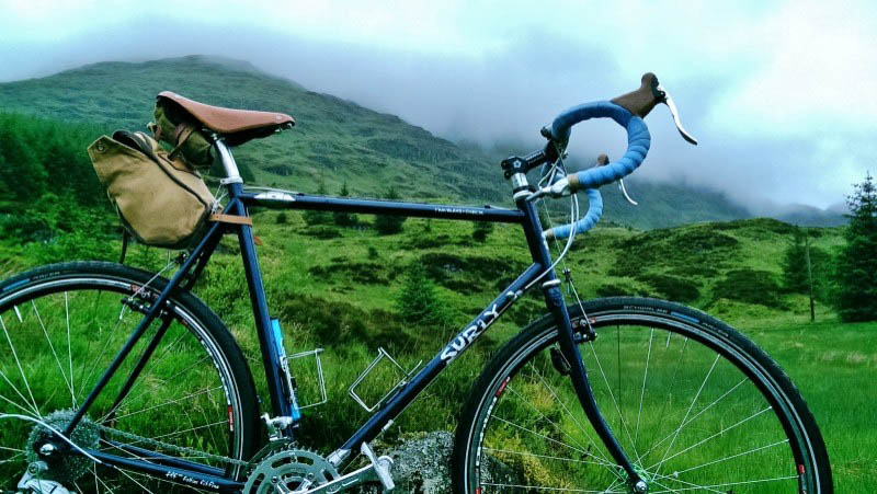 Right side view of a black Surly bike, with a green grass, mountain hill in the background with fog on top