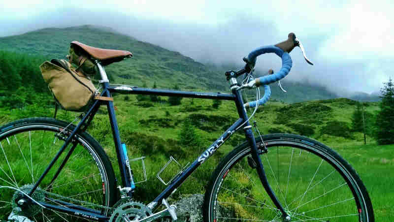 Right side view of a Surly bike, dark blue, with a green grass hill and fog above, in the background