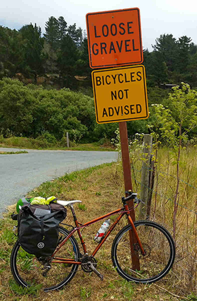 Right side view of a Surly bike with rear saddlebags, parked against a sign showing, Loose Gravel Bicycles Not Advised