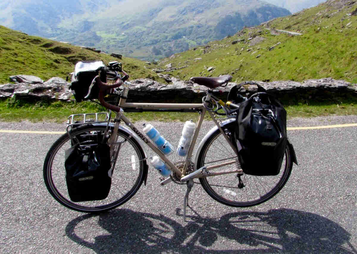 Left side view of a tan, Surly Long Hauler Trucker bike with gear, parked on a road, with a mountain valley behind